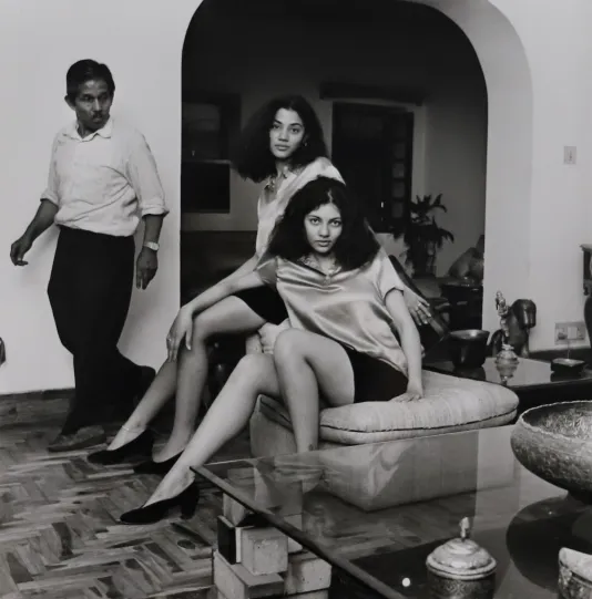 Blackened white photo of two women posing on a couch while a man walking by turns around to check them out.