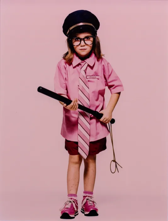 A young child wears a pink dress shirt and tie, pink sneakers, a police hat and has a baton in both hands against a pink backdrop.