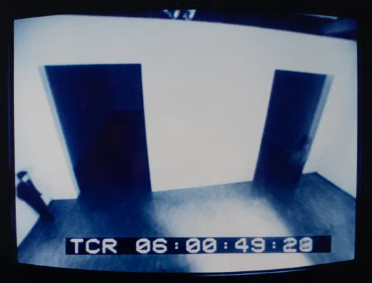 Screen capture from a security tape of a room with two large doorways and a person standing agains the left wall.