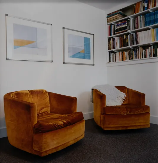 Photograph of an office featuring two plush, burnt orange swivel chairs with one white throw blanket, a bookshelf in the background and two prints hung on the wall.