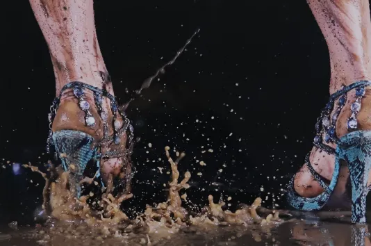 Close up photograph of a person wearing blue heels stepping in a brown puddle, making the water splatter all over.