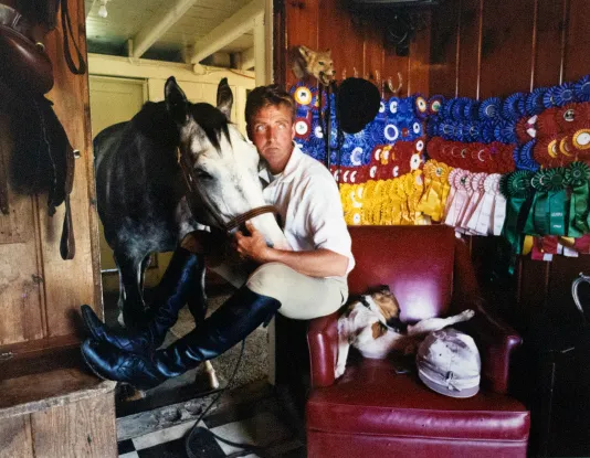 Man sitting on the arm of a red leather chair with a horse in a doorway resting its head on the man's lap.