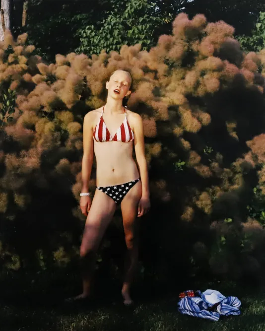 Photograph of a woman wearing an American flag bikini posing in front of a bush.