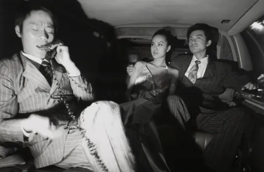 A couple sits in the back of a limousine while a man speaks on the phone. The woman looks at the man on the phone.