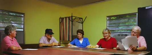 Town council meeting where three men and two woman sit at a wooden table with papers in their hands in a yellow room.
