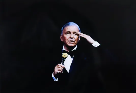 Portrait of Frank Sinatra with his left hand on his forehead and his right hand holding a microphone.