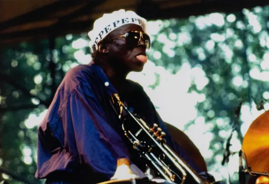 Man wearing a purple blouse holding an instrument with his tongue out.