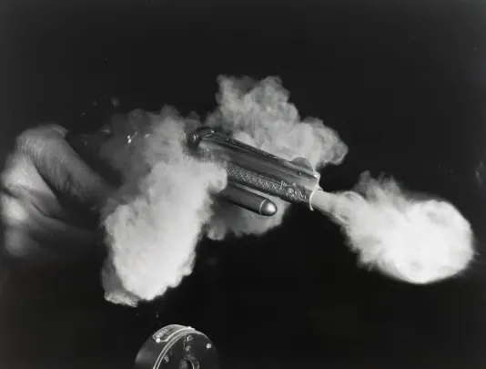 Black and white photograph of an antique gun mid fire with smoke coming out of the front and sides of the gun.