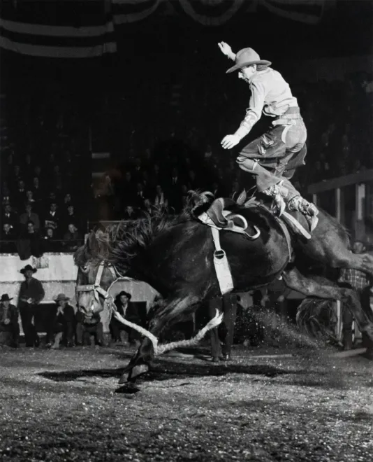 Black and white photograph of a cowboy jumping on top of a horse that has its front legs off the ground.