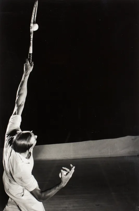 Black and white photograph of a man striking a tennis ball with his racket above his head.