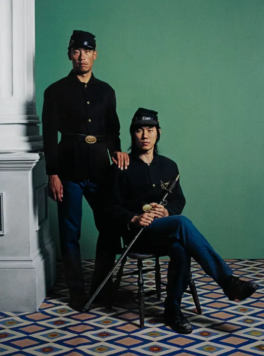 One man sits in a chair holding a sword and another man stand behind him with his right hand on the man's shoulder. There is a green wall behind them and a white pillar to the left.