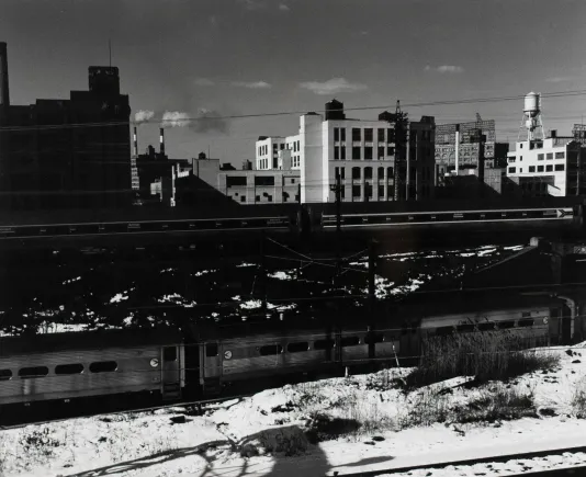 Black and white photograph of buildings in the background, a train in the middle ground and snow on the train platform in the foreground.