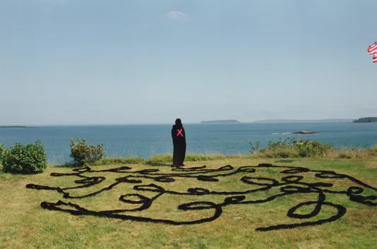 A person wearing all black with a large red X on their back stands on a grassy cliff that overlooks the ocean. The grass has a series of dark ropes in loops.