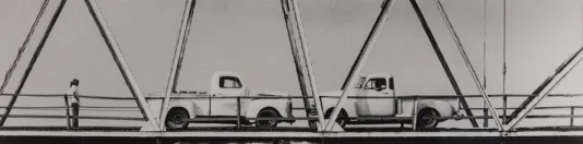 Two old fashioned pickup trucks on a suspension bridge while a person looks at them on the left.