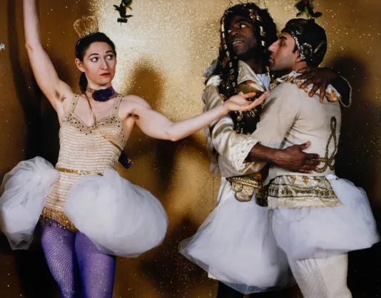 Three people in ballerina costumes stand in front of a gold backdrop. Two of them embrace each other and the third has her arms wide open and glances at the other two.