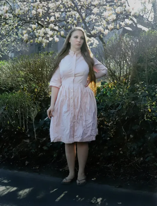 Photograph of a woman in a pink dress with a cigarette in her right hand posing in front a blossoming tree.