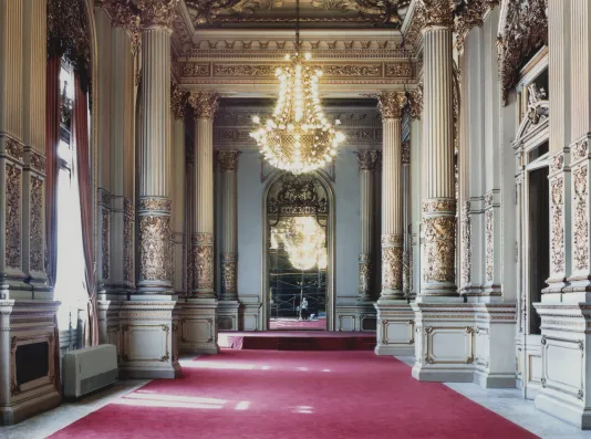Photograph of an empty hall featuring a lit chandelier and red carpets.