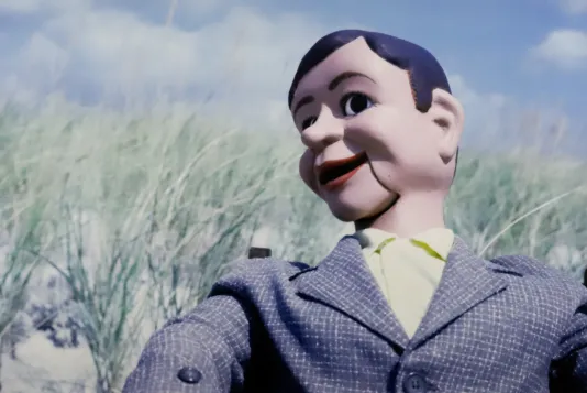 Photograph of a male dummy on the beach with sea grass and a cloudy blue sky in the background.