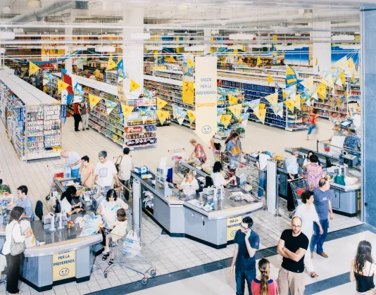 Photograph taken from up high busy checkout aisles at a supermarket featuring yellow flags hanging on the ceiling and the store aisles in the background.
