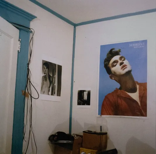 Photograph of a bedroom corner with white walls and teal trim and a large poster of Morrissey on the right wall and a black and white photo of a shirtless man on the left  wall.