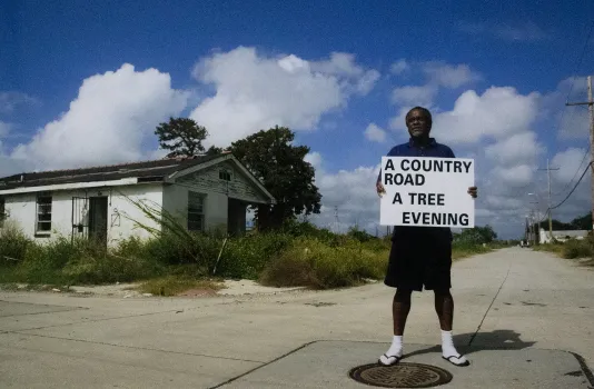 Photograph of a man in front of an abandoned, overgrown house holding a white sign hat reads "A country road a tree evening"