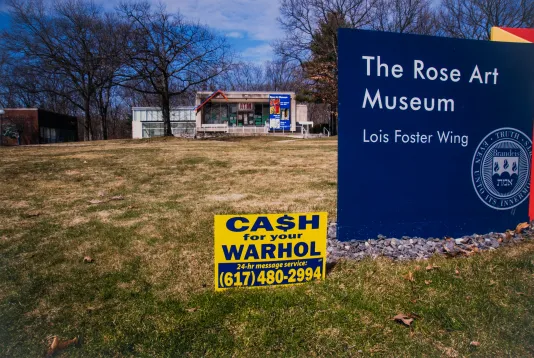 Photograph of the Rose Art Museum blue sign on a front lawn with a small yellow lawn sign that reads Cash for your Warhol with a phone number.