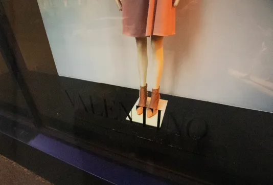 Photograph of a store window with a mannequin from the thighs down wearing a pink dress and brown boots. The glass reads Valentino.