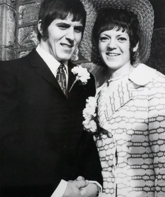 A man in a suit stands next to a woman in a dress with a fancy hat and they smile at the camera.