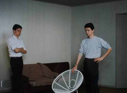 Double portrait of a man standing with his hand on a white chair and to the left, he stands with his arms crossed looking at the camera.