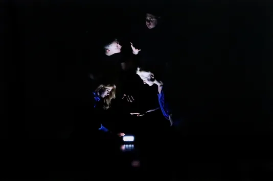 Dark image with a few blocks of phone light shining through in the center showing four people huddled together on their phones.