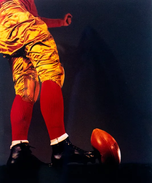 Photograph of a person from the waist down in red socks and gold football uniform about to kick a football.