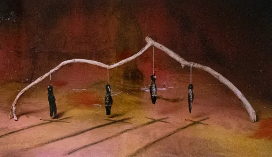 Red background with a large branch and four figures handing from the branch. In front of them, there are four tall shadows that resembles crosses.