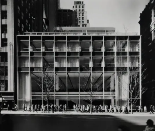Black and white photograph of a building with tall paneled windows and a long, straight line of people walking out front.