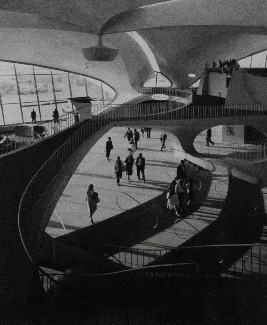 Black and white photograph of an airport terminal featuring a winding staircase and people walking.