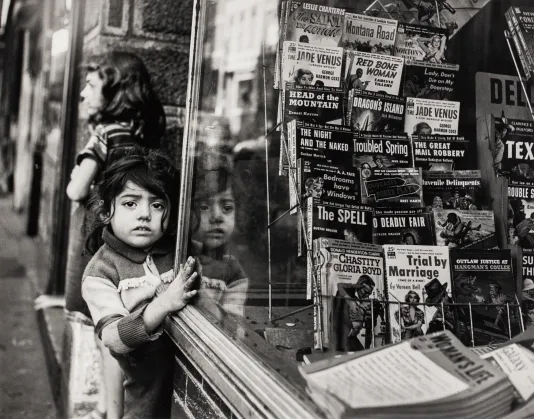 Black and white photograph of a child standing near a magazine stand holding onto the glass.