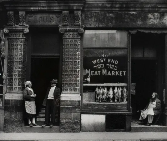 Black and white photograph outside a meat market with the words West End Meat Market written on the window and three people standing outside.