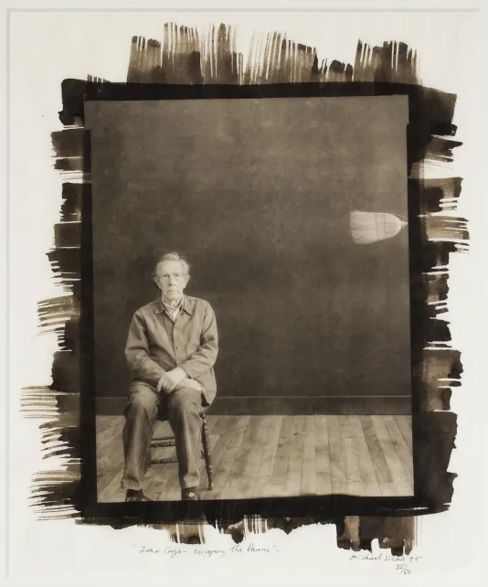 Black and white photograph with dark painted edge of a man sitting on a chair with a broom poking out on the right wall.