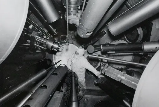 Black and white photograph of a person in a white space suit and a bunch of pipes all leading to where the person is standing.