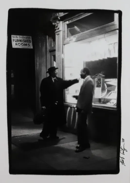 Black and white image of two men chatting outside a ticket booth on a street sidewalk.