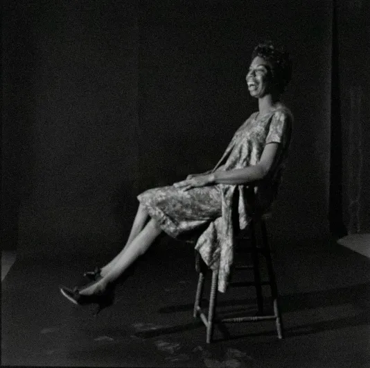 Black and white portraits of Nina Simone sitting in a chair with a dress and heels on smiling widely.