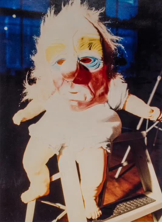 Baby doll with a red, blue, and yellow mask on its head sitting on the top of a step stool.