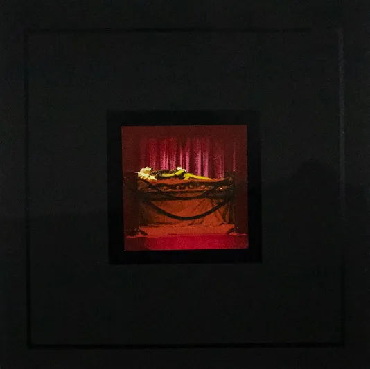 Dark image of a roped off red lit window and a person lying on a red bed.