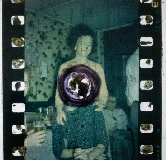 FIlm negative of a woman standing behind a seated woman with her hands on her shoulders. The seated woman's face is crossed out aggressively.