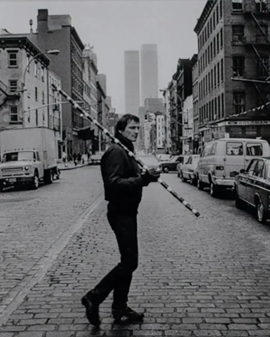 Black and white image of a man in the middle of a New York City street holding a striped pole surrounded by buildings.