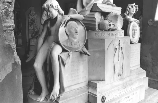 Black and white photograph of a marble sculpture featuring a figure sitting on the edge of a block holding a round plate with a man's face on it.
