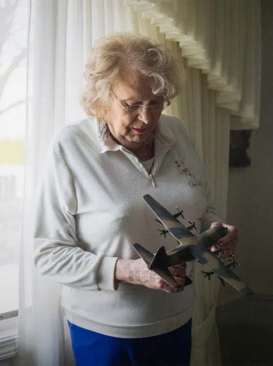 Older woman with white hair wearing a white sweater looks down at a model army airplane she is holding.