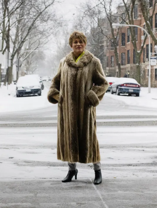 Woman in a long fur coat poses on a snow covered city street.