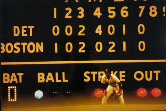 Red Sox baseball player on a field with a jumbo score board behind him.