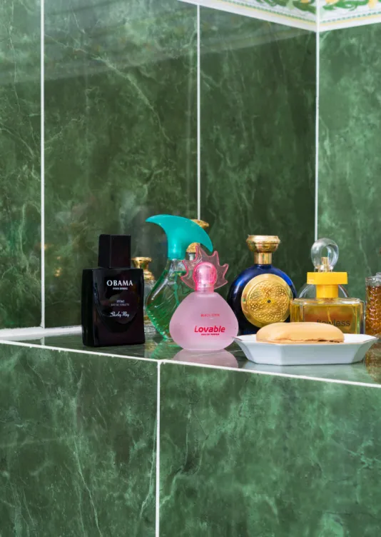 Bathroom shelf of different perfume bottles against a dark green tiled wall.