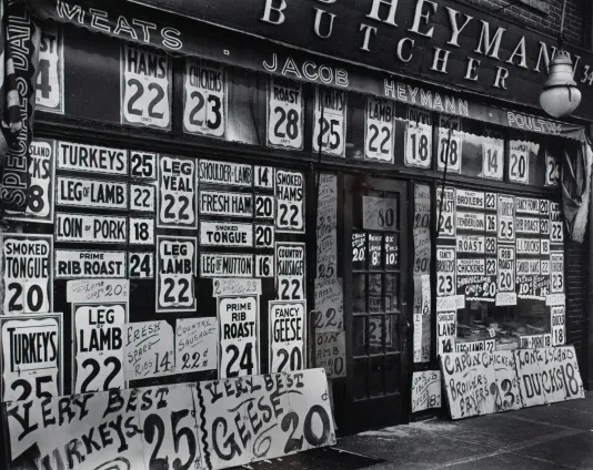 Black and white photograph of a butcher shop store front with the front covered in signs that have prices.